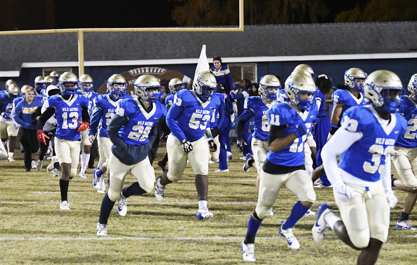 Football players in blue and gold uniforms run onto the field during a night game, with coaches and spectators visible in the background.