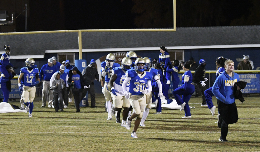 Football players in blue and gold uniforms run onto the field at night, followed by coaches and teammates, with a goalpost and spectators in the background.