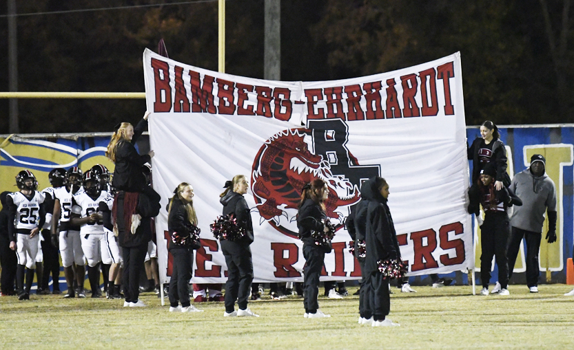 Cheerleaders stand in front of a large banner reading "Bamberg-Ehrhardt Red Raiders" on a football field, preparing for a sports event.