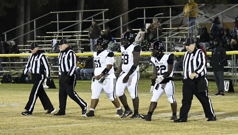 Football players in white uniforms and a referee walk across a field at night, with bleachers and spectators in the background.