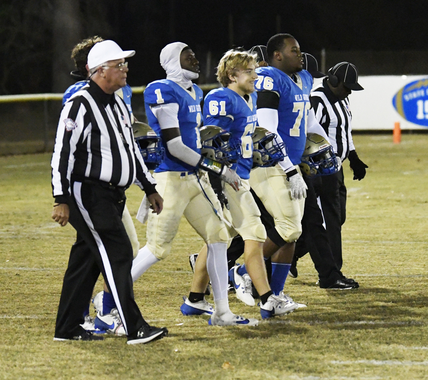 Football players and referees walk together onto the field at night, wearing uniforms and preparing for the start of a game.