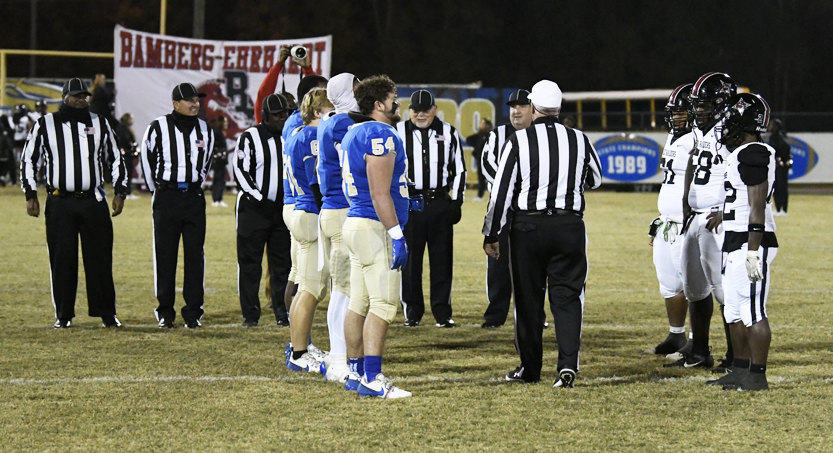 Football players in blue uniforms and referees in black and white stripes stand on a grass field during a nighttime coin toss before a game.