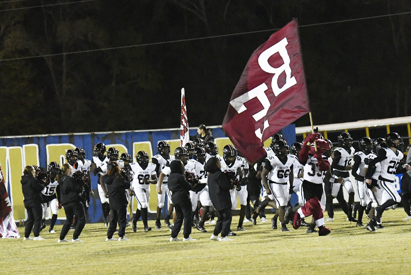 A football team in black and white uniforms runs onto the field, with one player holding a large maroon flag displaying bold white letters.