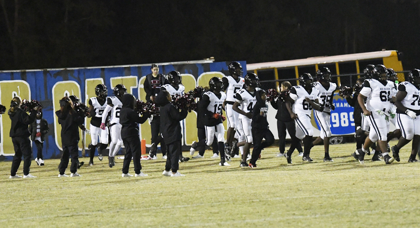 A football team in black and white uniforms runs onto a field at night, with coaches and players celebrating near a blue and yellow sign.
