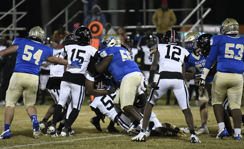 Football players in black and white uniforms tackle a player in blue and gold during a nighttime game on a grassy field.