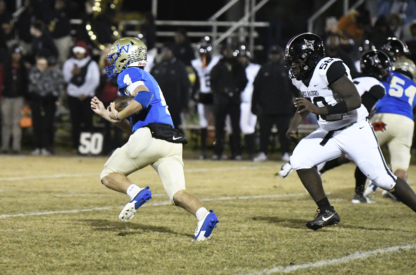 A football player in blue runs with the ball as a defender in white and black chases him on a grassy field during a game.