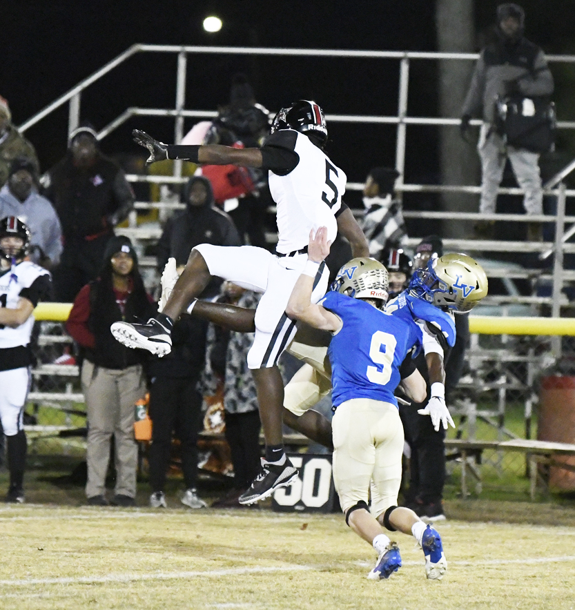 A football player in a white uniform jumps to catch the ball while a player in blue tries to defend, with spectators in the background.