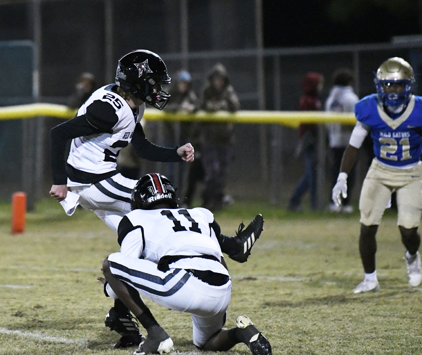 A football player kicks a field goal as a teammate holds the ball, while opposing players watch on a grassy field during a night game.