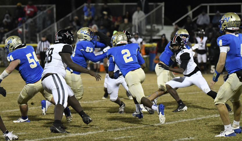 High school football players in blue and white uniforms compete on a grassy field at night, with spectators watching from bleachers in the background.