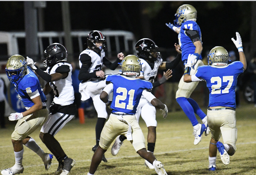 Football players in blue and white uniforms compete against players in black and white uniforms under stadium lights during a game.