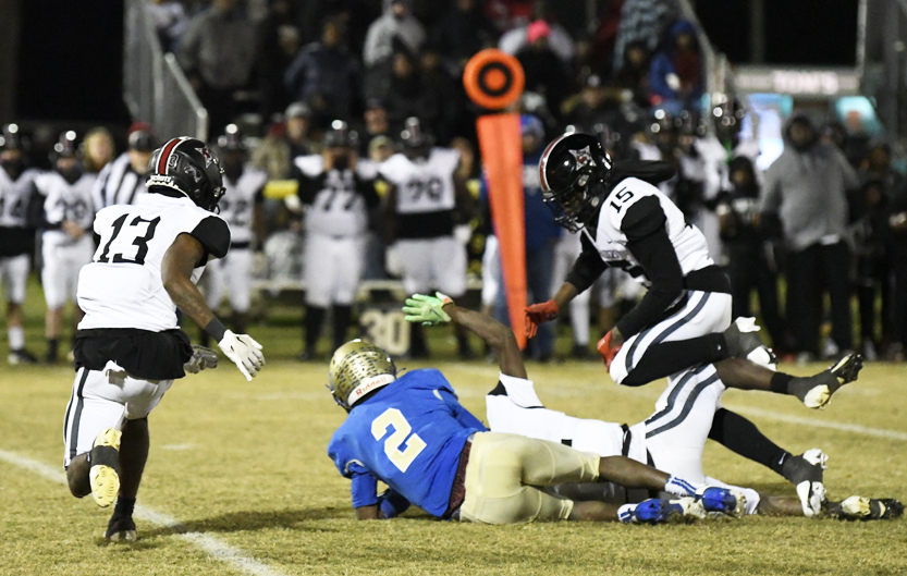 Football players in action on the field at night, with one player in blue tackled by opponents in black and white uniforms, and a crowd watching in the background.