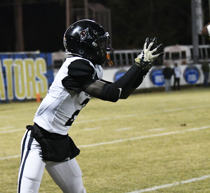 A football player in a black helmet and white uniform extends his arms, preparing to catch a pass on a lit outdoor field at night.