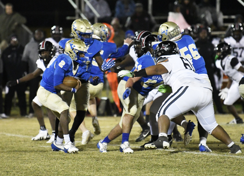 High school football players in blue and gold uniforms play against a team in white uniforms during a nighttime game on a grassy field.