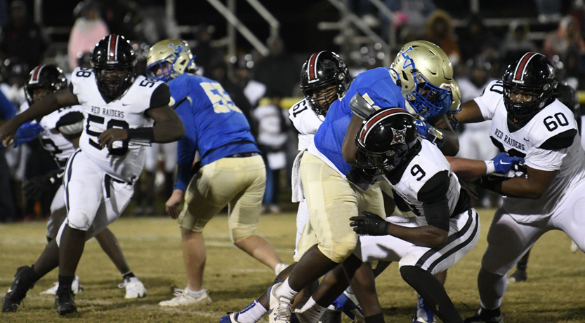 High school football players in blue and white uniforms compete on a grassy field during a night game, with action focused on blocking and tackling.