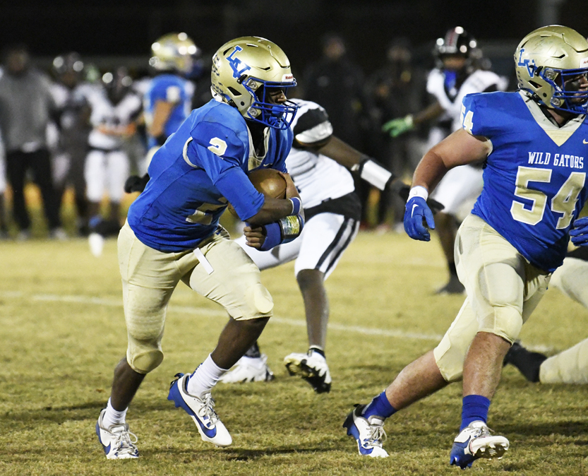 Football player in blue and gold uniform carrying the ball during a nighttime game, with teammates and opponents visible in the background.