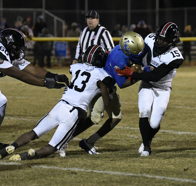 Three football players in white uniforms tackle a player in blue, while a referee watches in the background on a grassy field at night.