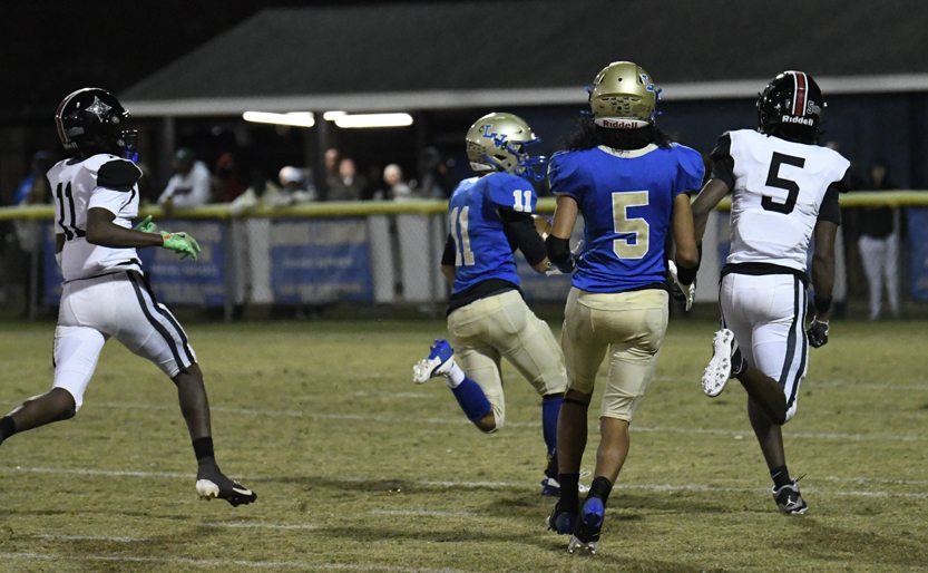 Football players in blue and gold uniforms run on a grass field during a nighttime game, with spectators watching from the background.