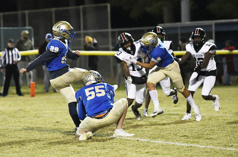 High school football players in blue and gold uniforms attempt a field goal as opponents in black and white try to block the kick during a night game.