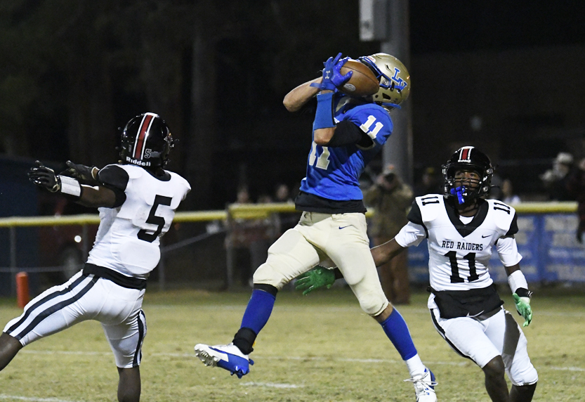 A football player in blue catches a pass mid-air while two opponents in white uniforms attempt to defend during a night game.