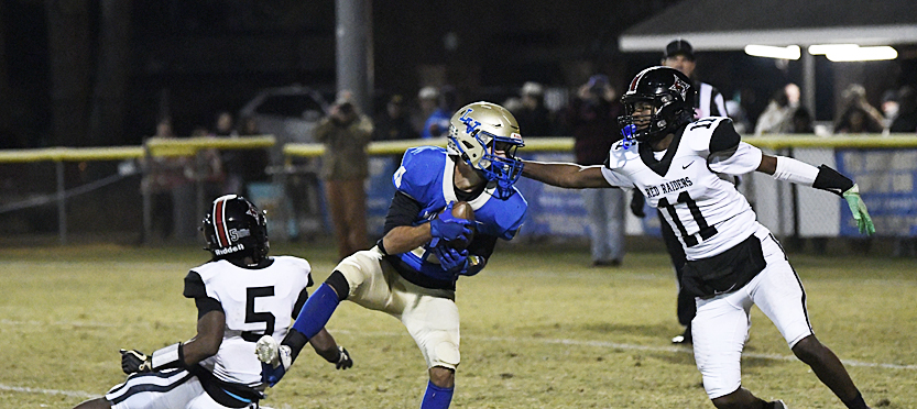 A football player in blue catches a pass while another player in white attempts to block him during a nighttime game.