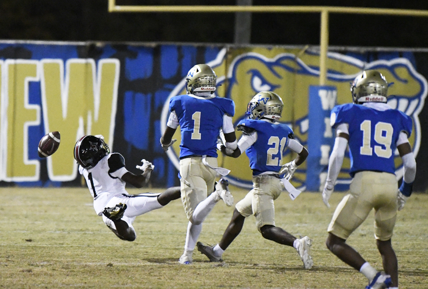 A football player in white is falling as three players in blue run past on a field with a painted backdrop and goalpost.
