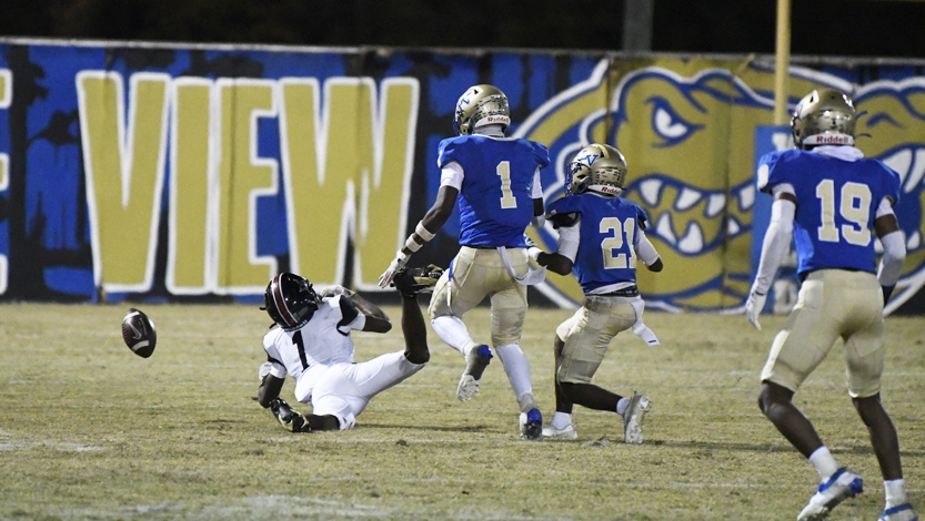 Three high school football players, two in blue and gold uniforms and one in white, compete during a night game on a grassy field with a colorful mascot banner in background.