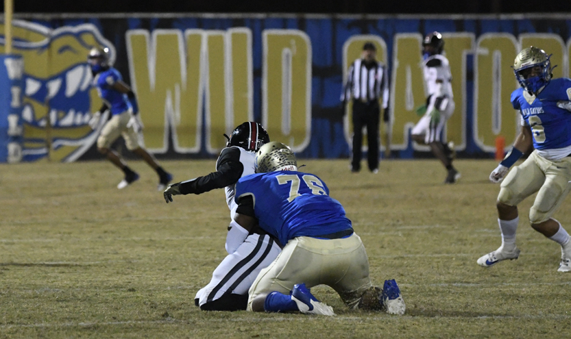 A football player in blue and gold tackles a player in white and black on a field with a "Wildcats" banner in the background.