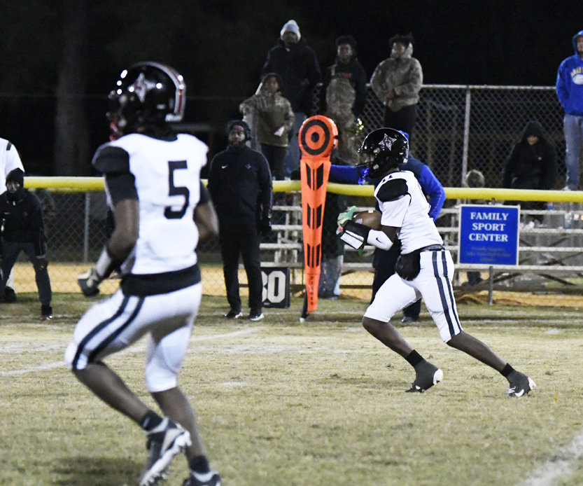 Two American football players in white uniforms on a field at night, with one player running and the other preparing to throw the ball. Spectators watch from behind the fence.