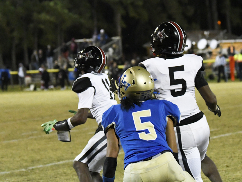 Three football players on the field, two in white uniforms and one in a blue uniform, all wearing helmets during a night game.