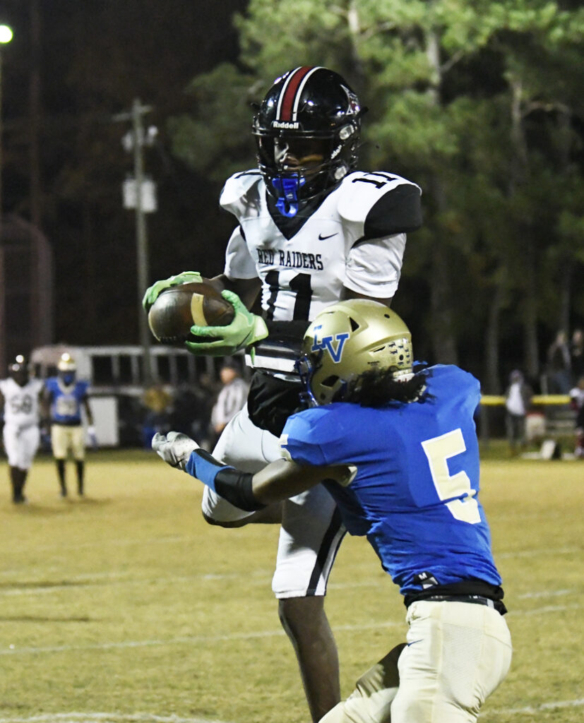 A football player in black and white catches a pass above a defender in blue during a nighttime game on a grassy field.