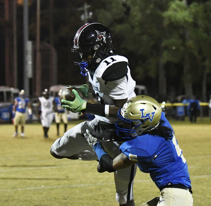 Football player in a white uniform jumps to catch a pass while being tackled by an opponent in a blue and gold uniform on a grassy field.