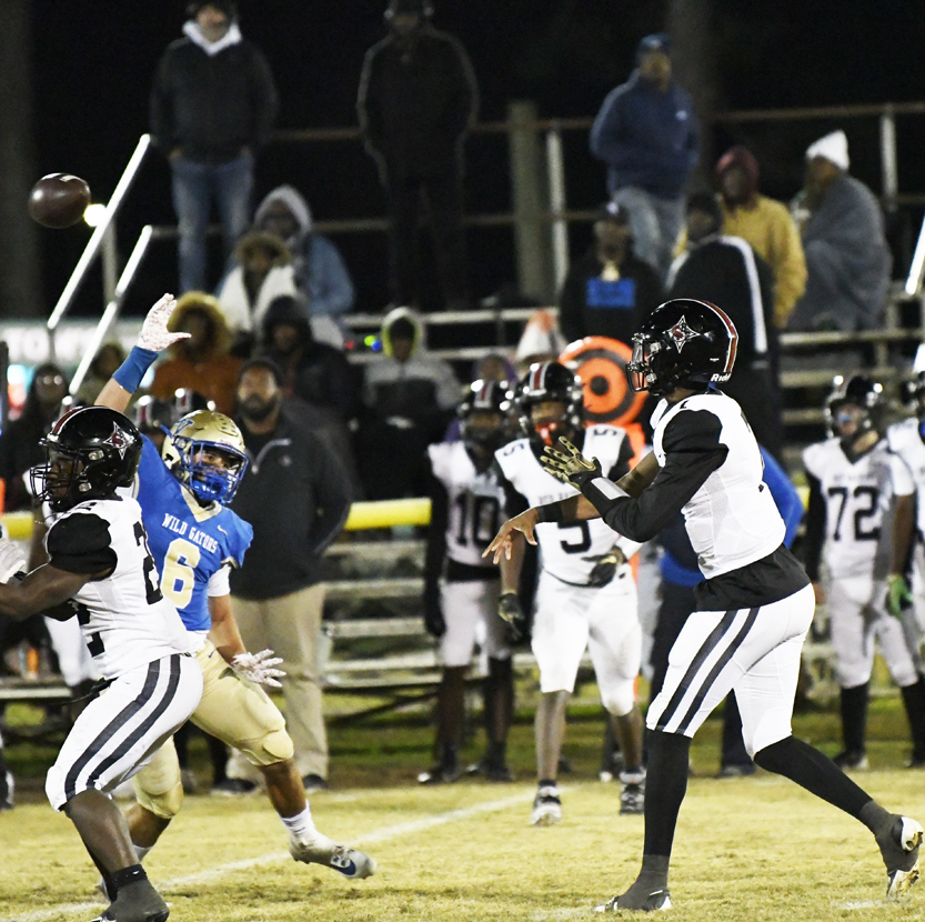 A football player in black prepares to throw the ball while players in blue and white attempt to block during a nighttime game.