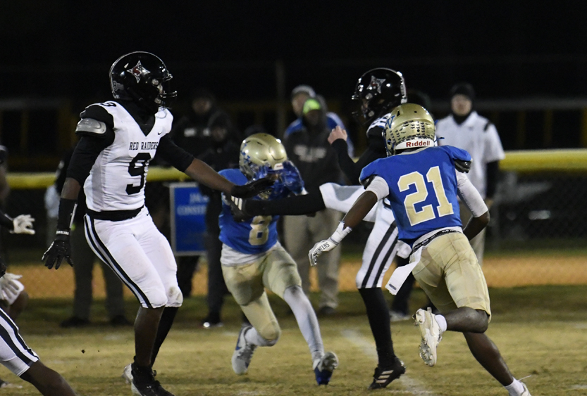 High school football players in blue and white uniforms compete on a field at night, with coaches and spectators visible in the background.