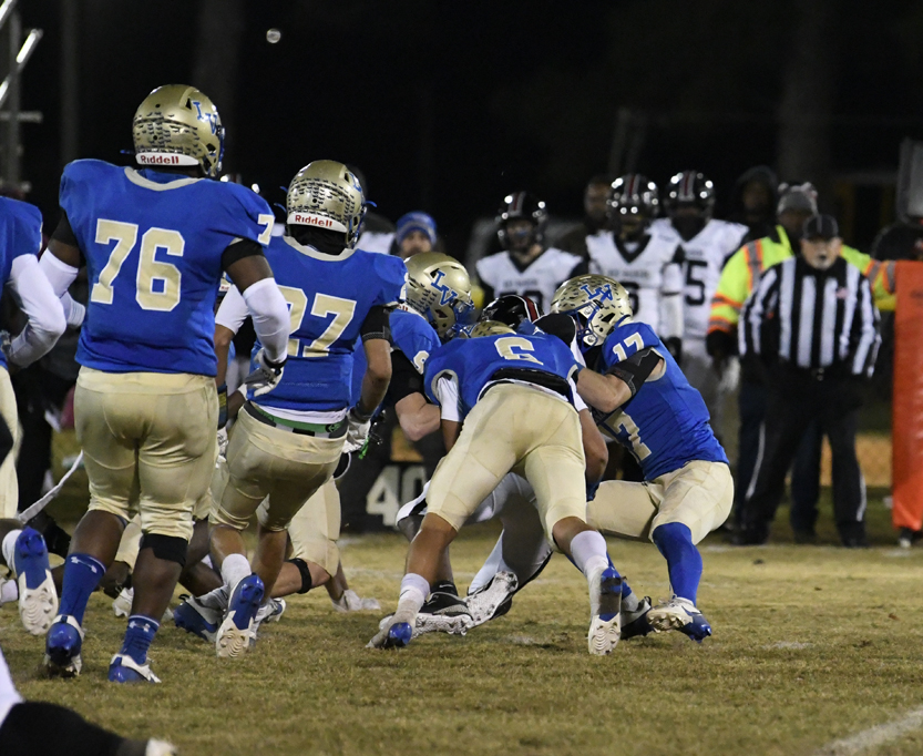 Football players in blue and gold uniforms tackle an opponent on a grassy field at night, with referees and team members watching in the background.