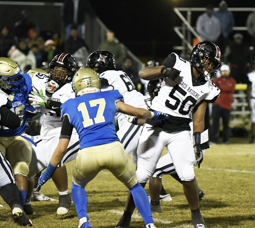 Football players in blue and white uniforms compete during a night game, with one player wearing number 17 facing the opposing team. Spectators watch from the stands.
