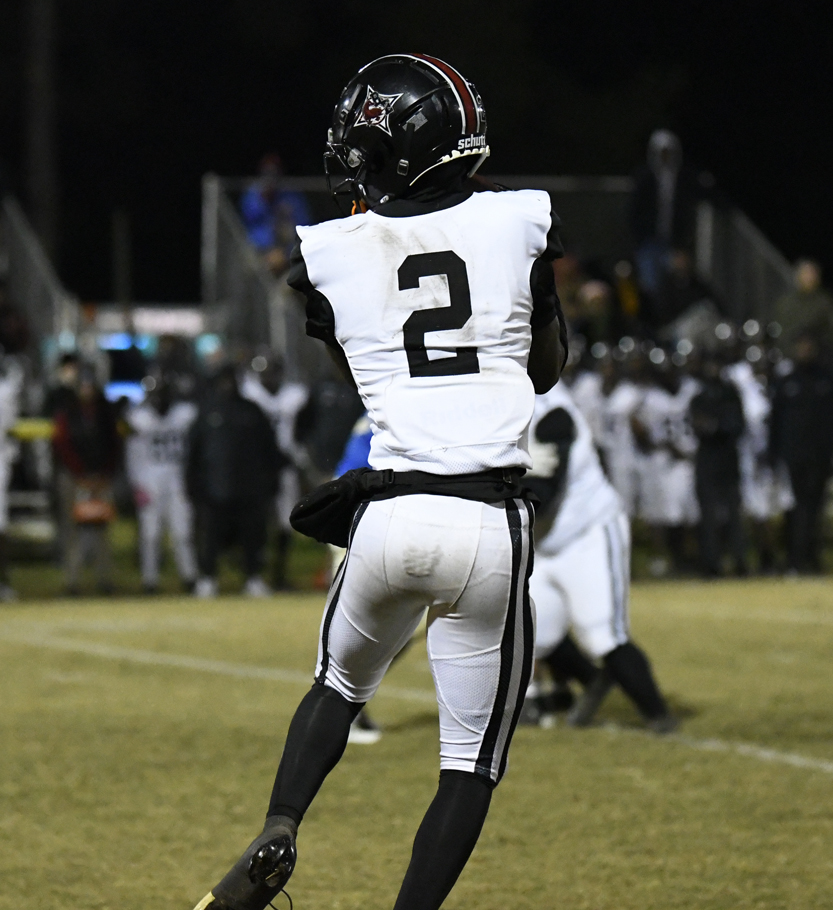A football player wearing a white uniform with the number 2 catches a pass during a night game, with teammates and spectators visible in the background.