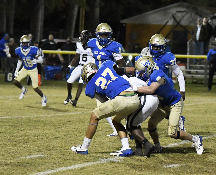 Football players in blue and gold uniforms tackle an opponent in white during a nighttime game on a grassy field.