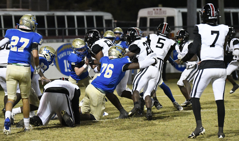 Football players in blue and white uniforms tackle each other during a night game on a grassy field, with school buses visible in the background.