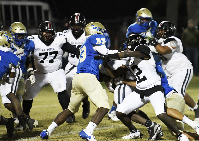 High school football players in blue and white uniforms engage in a play during a nighttime game on a grassy field.