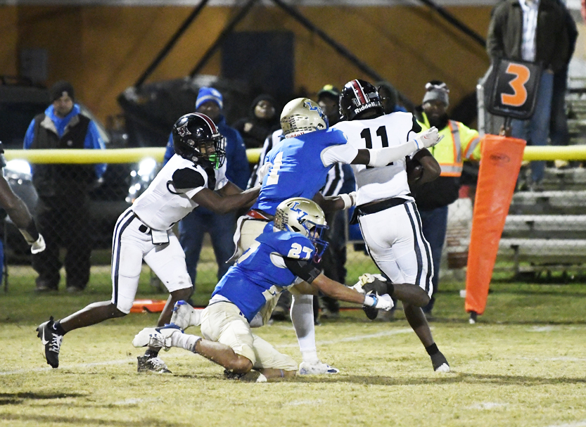 Football players in blue and white uniforms compete on a field at night, with one player tackling another as officials watch from the sidelines.