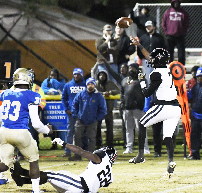 Football players in blue and white uniforms compete during a nighttime game, with one player jumping to catch the ball as spectators watch from the sidelines.
