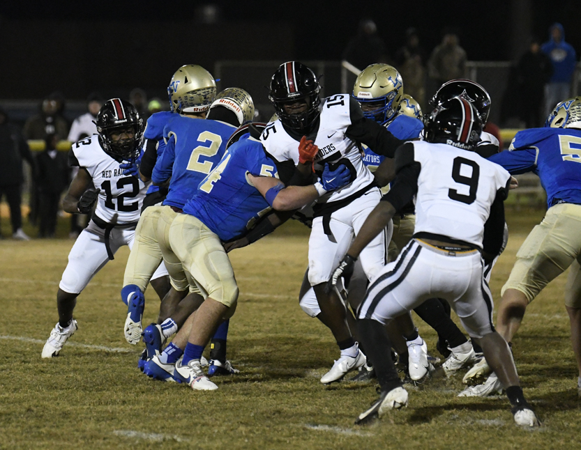 High school football game at night, players in blue and white uniforms collide on the field during an active play.