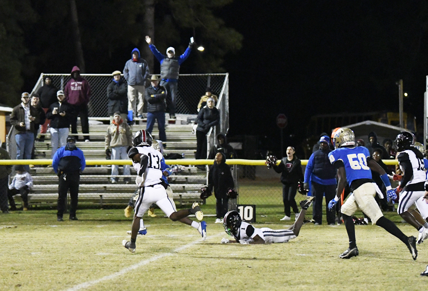 Football players in action on a field at night, with one player falling and spectators cheering from bleachers in the background.