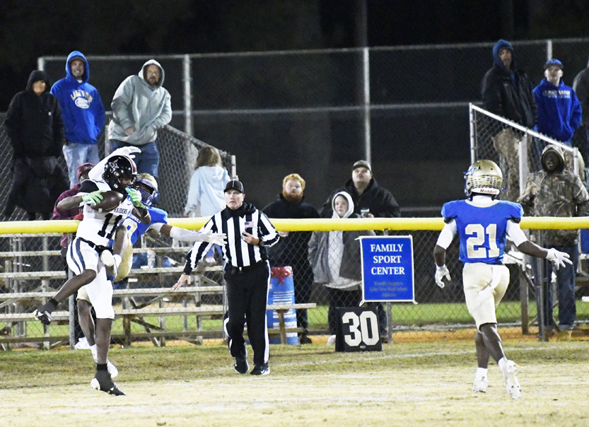 Football players in action near the sideline, with a referee watching, spectators in the bleachers, and a blue sign and scoreboard visible in the background.