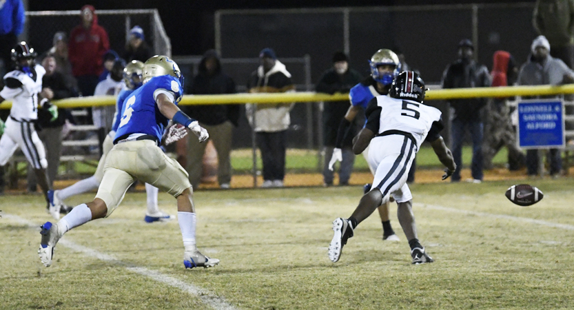 Players in blue and white football uniforms compete on a field at night, with spectators bundled in coats behind a yellow barrier.