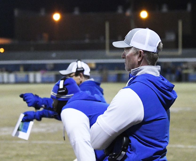 Football coaches in blue and white gear stand on the sideline during a nighttime game, with lights glowing in the background and playbooks in hand.