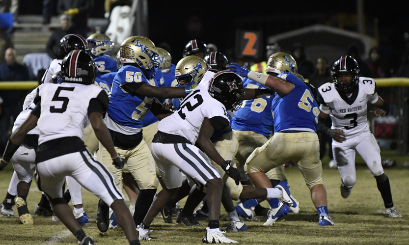 Football players in blue and gold uniforms block opponents in white and black uniforms during a night game on a grassy field.