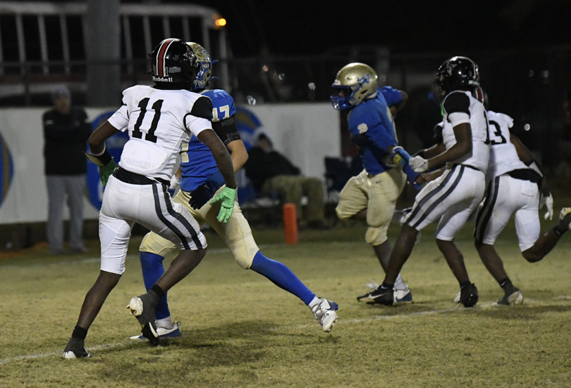 Football players in blue and white uniforms compete under stadium lights on a grassy field during a night game.