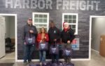 Four people stand in front of a Harbor Freight sign, each holding bags, with more bags arranged in front of them indoors.