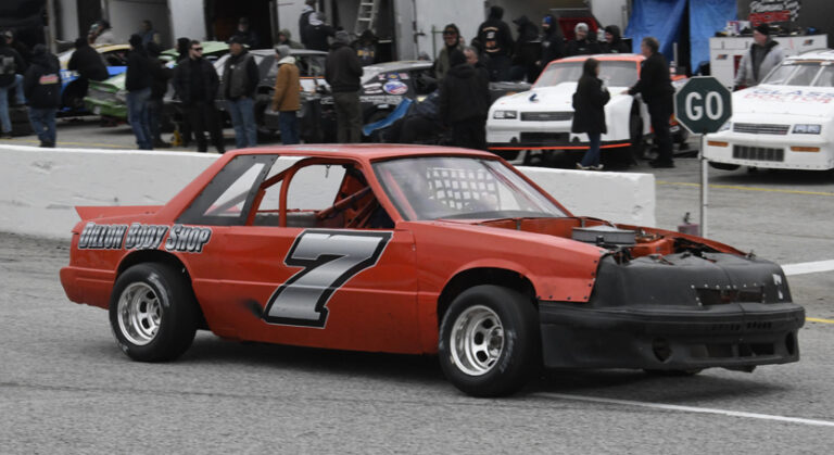 A red race car with the number 7 on the side is parked on a track, surrounded by people and other vehicles in the background.